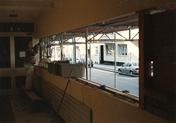 Blick auf eine Baustelle im Jugendhaus Düsseldorf, 1997. Ein Handwerker arbeitet an einem Fensterrahmen, während Gerüste sichtbar sind. Auf der Straße parken mehrere Autos.