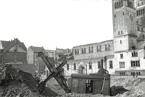Baustelle des Jugendhauses Düsseldorf mit einem Bagger, der in einem Erdhaufen steht. Im Hintergrund sind historische Gebäude und ein Kirchturm sichtbar.