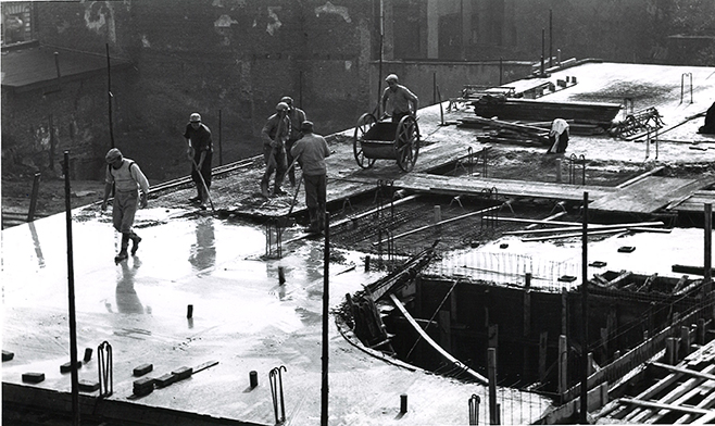Sechs Arbeiter auf einer Baustelle in Düsseldorf, die Beton auf einer Fläche gießen. Einige nutzen Schaufeln, andere einen Karren. Baustellenmaterialien sind sichtbar.